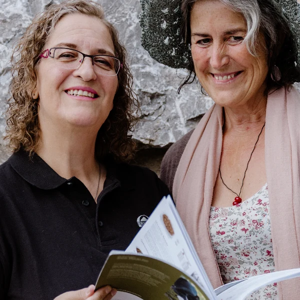 Zwei Frauen stehen im Freien vor einer Steinmauer, lächeln und schauen auf ein aufgeschlagenes Buch, das eine von ihnen in der Hand hält.